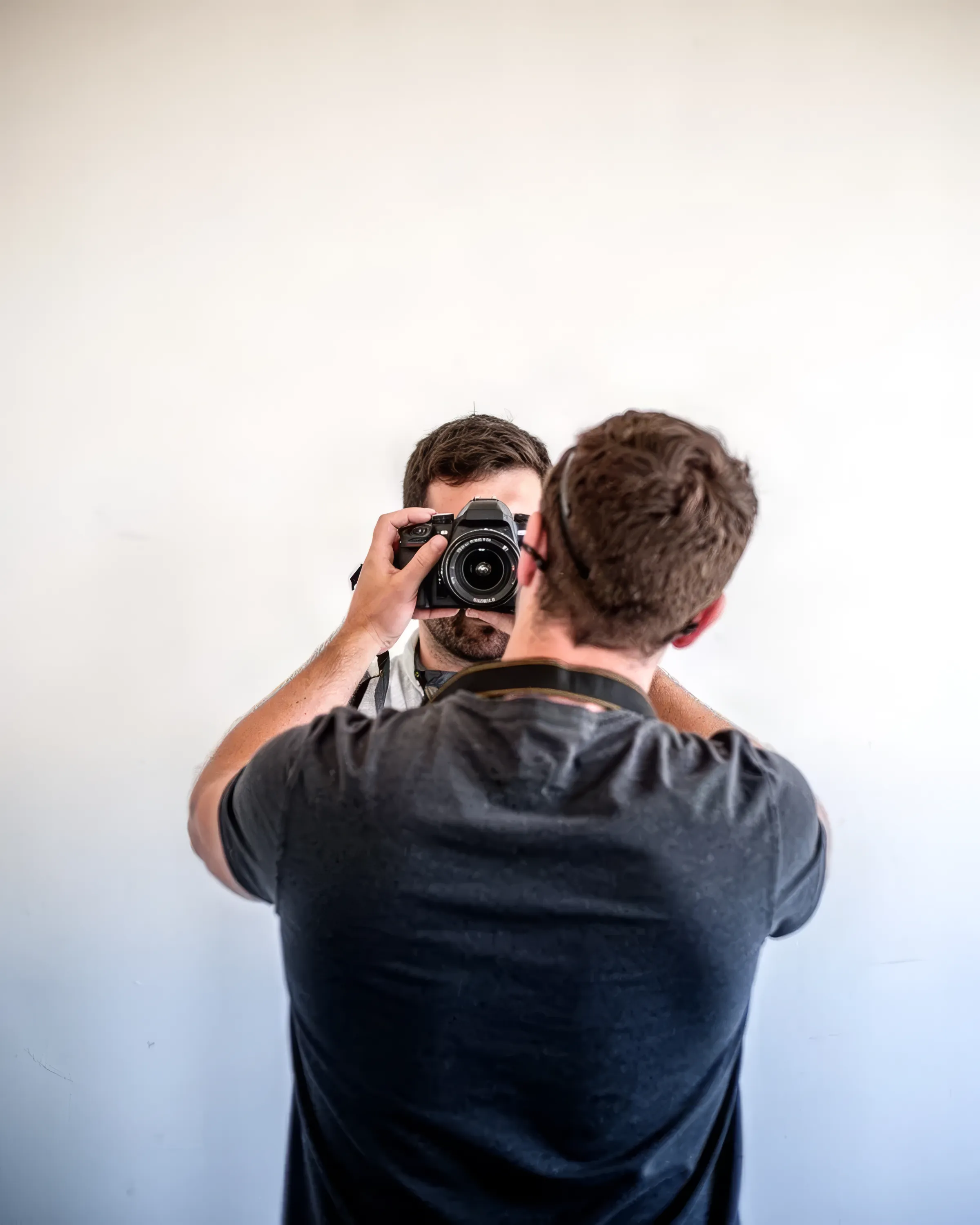Ein Fotograf steht von hinten vor einer hellen Wand; im Spiegel hebt er die Kamera vors Gesicht.