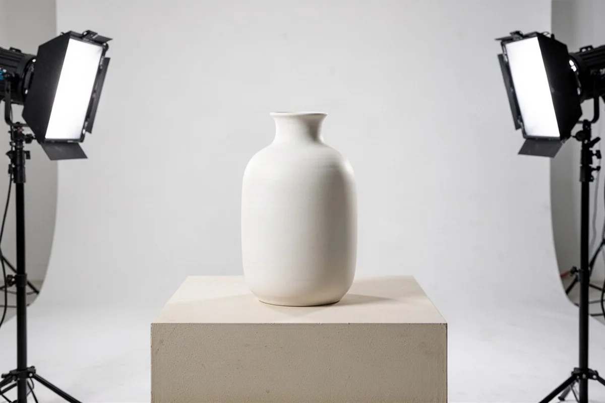 Eine cremeweiße Vase steht auf einem Podest zwischen zwei Flächenleuchten vor hellem Hintergrund.