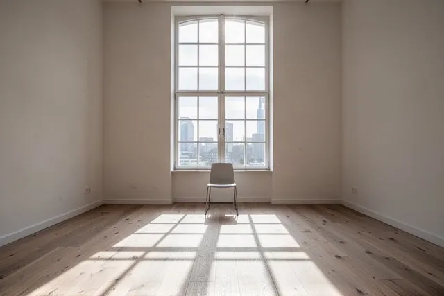 Heller Büroraum mit hohem Sprossenfenster, Morgensonne auf dem Holzdielenboden, ein einzelner Stuhl.