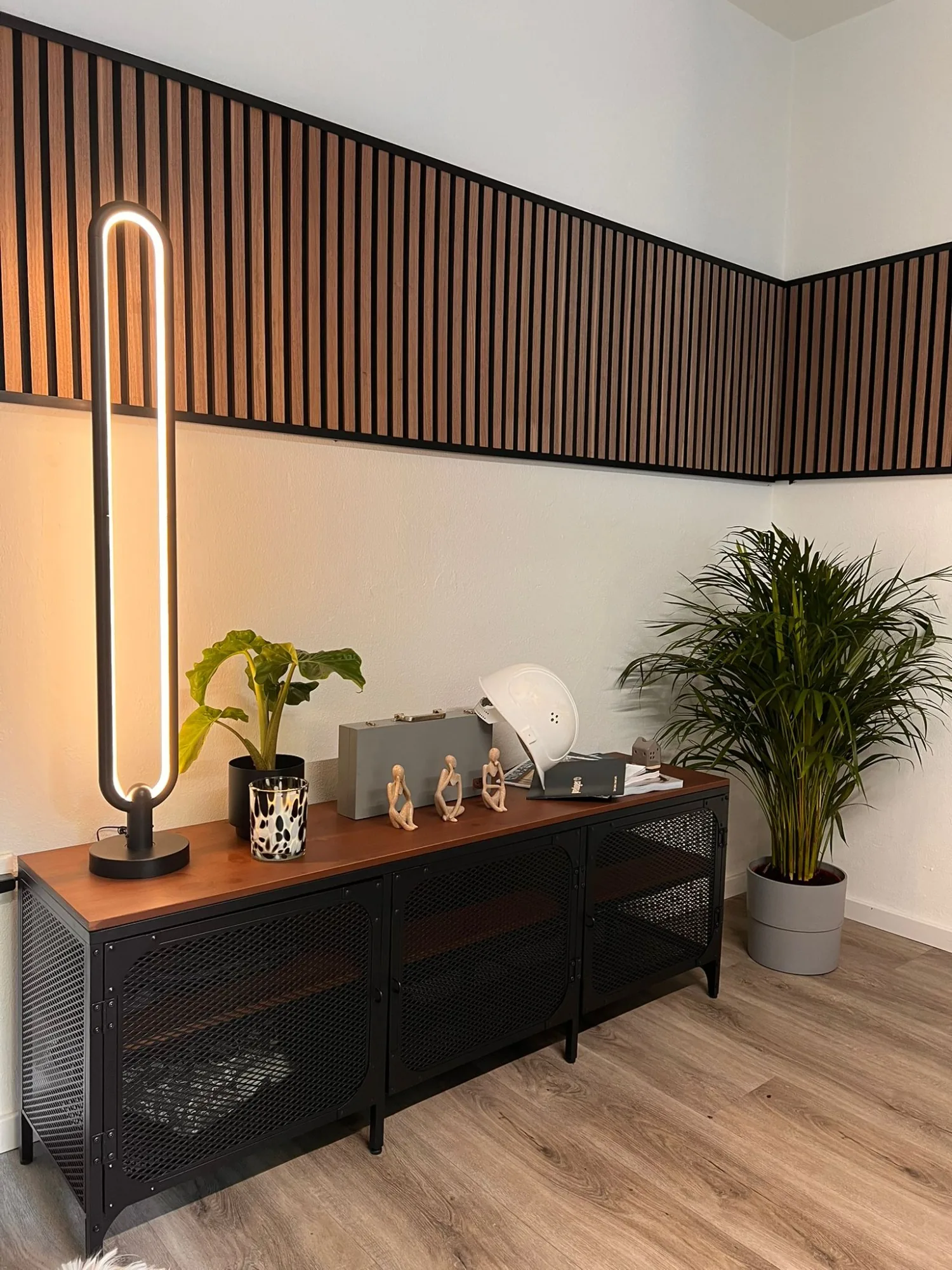 Sideboard im Büro vor einer Wand aus schwarzen Holzlamellen, daneben Stehleuchte und Pflanze