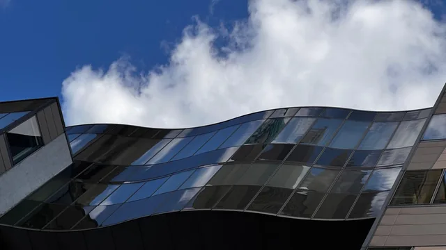 Gewellte dunkle Glasfassade eines großen Gebäudes, in der sich Wolken spiegeln
